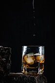 Whiskey drops falling on ice cubes served in crystal glass placed on rough surface against black background