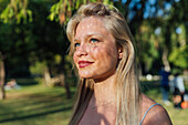 Glückliche Frau mit blondem Haar und Schatten im Gesicht, die an einem sonnigen Sommertag in einem Park steht und wegschaut