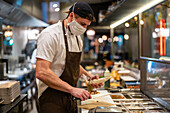 Focused professional male cook in uniform and protective mask standing with knife and pastry bag while working in restaurant at coronavirus epidemic