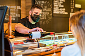 Concentrated barman in protective mask and apron standing at counter and pouring red wine into glasses for unrecognizable woman customer in cafe during coronavirus