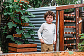 Lockig behaarter Junge steht neben einem Gartenbeet mit gepflanzten Sprossen im Garten und schaut in die Kamera