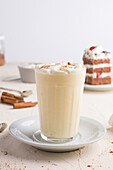 Glas Milchpunsch mit Zimtpulver auf geschlagenem Eiweiß vor Kuchenstück auf Cafeteria-Tisch vor hellem Hintergrund