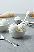 Vanilleeis in weißen Keramikschalen mit Löffeln neben Waffeltüten auf einem grauen Tisch