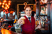 Smiling female barkeeper standing at bar counter with a type of alcohol drink served in creative cocktail glasses in shape of mushroom