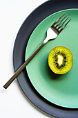 From above of half of fresh ripe juicy kiwi and fork placed on grey and green plates on white surface