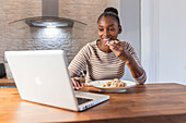 Afroamerikanerin genießt leckeren Patacon mit Topping, während sie mit ihrem Netbook in der Küche zu Hause im Internet surft