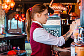 Seitenansicht einer Barkeeperin bei der Arbeit in der Bar, die die Kasse bedient und den Bildschirm des Displays berührt