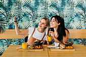 Young cheerful best female friends taking self portrait on cellphone during brunch in cafe