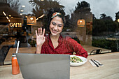 Through window of cheerful ethnic female sitting at table with healthy food in restaurant and waving hand at laptop screen during online meeting with friends