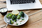 Yummy cooked broccoli with lemon slice and cashew nuts near bowl with white sauce and netbook on wooden table