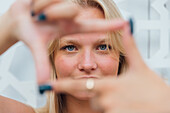 Zufriedene Frau mit blondem Haar, die ein Rahmungszeichen zeigt und durch die Finger in die Kamera schaut