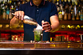 Unrecognizable bartender pouring crushed ice to the glass while preparing mojito cocktail in the bar