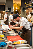 Side view of professional male cook in protective mask adding cream from pastry bag while preparing delicious burger in restaurant kitchen at work with colleagues during coronavirus epidemic