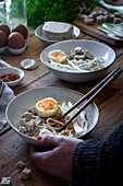 Von oben abgeschnittene, nicht erkennbare Person isst frisch gekochte Ramen-Nudeln mit Tofu, Eiern und Gemüse mit Stäbchen auf einem Holztisch