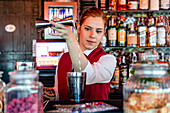 Fokussierte weibliche Barkeeperin in Uniform gießt alkoholisches Getränk aus einem Jigger in einen Shaker, während sie einen Cocktail in einer Bar zubereitet