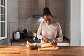Black female in apparel with striped ornament chopping cooking banana with knife on cutting board at table in house