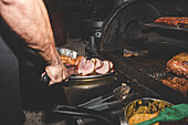 Crop anonymous male cook in tongs serving grilled pork on plate while standing near modern barbecue during work in cafe