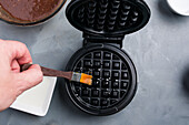 From above of crop unrecognizable person using brush to spread butter on black waffle maker at gray table in kitchen