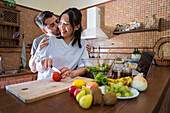 Husband kissing happy Asian wife cutting tomato for healthy salad during lunch preparation in kitchen at home
