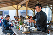 Seitenansicht eines ernsten Mannes mit Armbändern, der an einem Tisch auf der Terrasse stehend leckeres Essen aus einem Topf einschenkt