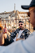 Erwachsener bärtiger Mann mit modischer Sonnenbrille sitzt auf einer Couch mit einer Flasche Bier in der Nähe eines Freundes auf einer Terrasse in Kappadokien, Türkei