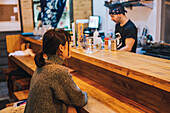 Side view of Asian woman in casual wear sitting at counter and talking with male worker of modern ramen bar