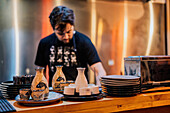 Junger Mann in Schürze kocht asiatische Gerichte, während er am Tresen einer Ramen-Bar steht
