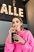 Fröhliche junge Frau in rosafarbenem Pullover nippt an einem kalten Erfrischungsgetränk durch einen Strohhalm, während sie ihre Freizeit in einer Cafeteria verbringt und fröhlich in die Kamera schaut