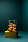 Front view of a glass cup with cold whiskey and cubes of ice placed on a gold-colored rugged base and a turquoise blue corner background