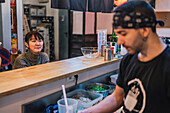 Asian woman in casual wear sitting at counter and talking with male worker of modern ramen bar