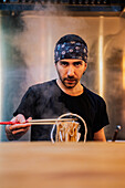 Young bearded male chef in bandana looking at camera while holding noodle with chopsticks in ramen bar
