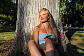 Unbeschwerte Frau, die mit ihrem Smartphone telefoniert und über Kopfhörer Musik hört, während sie im Sommer bei Sonnenuntergang unter einem Baum im Park sitzt und in die Kamera schaut