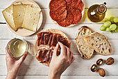 Top view crop anonymous female chef arranging delicious bacon slices on platter placed on table near plates with cheese and wurst cuts near olive oil and bread