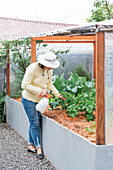 Ganzkörperansicht einer beschäftigten Gärtnerin mit Hut und Flasche, die grüne Pflanzen im Gartenbeet eines Bauernhofs besprüht