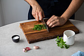Unrecognizable male in apron cutting fresh parsley on wooden cutting board near salt and garlic while cooking lunch at home