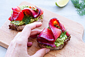 Cropped unrecognizable person hand holding tasty appetizers of rye bread toasts with avocado spread and smoked salmon slices with dill sprigs on chopping board