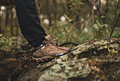 Crop unrecognizable person in trekking boots standing on mossy rock in forest in countryside