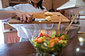 Anonymous woman putting cut banana from chopping bowl into bowl with healthy salad during lunch preparation at home