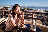 Glückliche Asiatin mit einem Glas Heißgetränk in der Hand, die wegschaut, während sie beim Frühstück auf dem Balkon an einem Tisch mit Essen sitzt