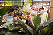 Weibliche Einkäuferin mittleren Alters mit einer Gesichtsmaske aus Stoff schaut weg, während sie auf eine Pflanze in einem Florarium in einem Gartencenter zeigt