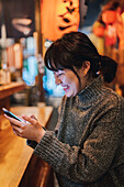 Asian lady in casual sweater smiling while using mobile phone at counter in traditional ramen bar