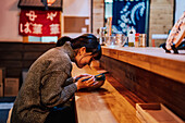 Seitenansicht einer zufriedenen Asiatin in Freizeitkleidung, die mit Stäbchen und einer Schüssel mit Ramen in einem Café am Tresen sitzt