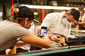 Waitress in uniform and protective mask taking photo on smartphone of chef and cooks preparing food in open kitchen during coronavirus outbreak