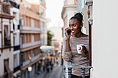 Afroamerikanerin mit einer Tasse Heißgetränk, die auf einem Balkon mit ihrem Handy telefoniert