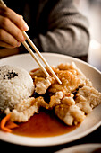 Hand einer Frau, die am Tisch sitzt und mit Stäbchen chinesisches gebratenes Fischgericht von einem weißen Keramikteller isst