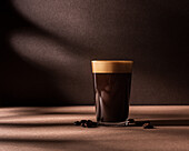 Vorderansicht einer schwarzen Kaffeetasse mit einer Schaumschicht, die neben einigen Kaffeebohnen auf einem braunen Tisch und einer Wand mit dunklen Schatten steht