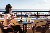 Happy Asian female with glass of hot drink in hand looking at camera while sitting on balcony at table with food during breakfast