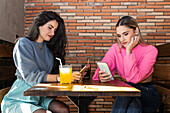 Junge stilvolle Freundinnen surfen auf ihren Handys, während sie in einer Cafeteria an einem Tisch mit Getränken sitzen