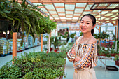 Satisfied young Asian female shopper with folded arms looking at camera against potted plants in garden store