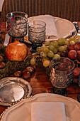 Kristallgläser in der Nähe von Teller und Besteck auf einem mit Weintrauben, Calluna vulgaris-Blüten und Granatäpfeln geschmückten Tisch